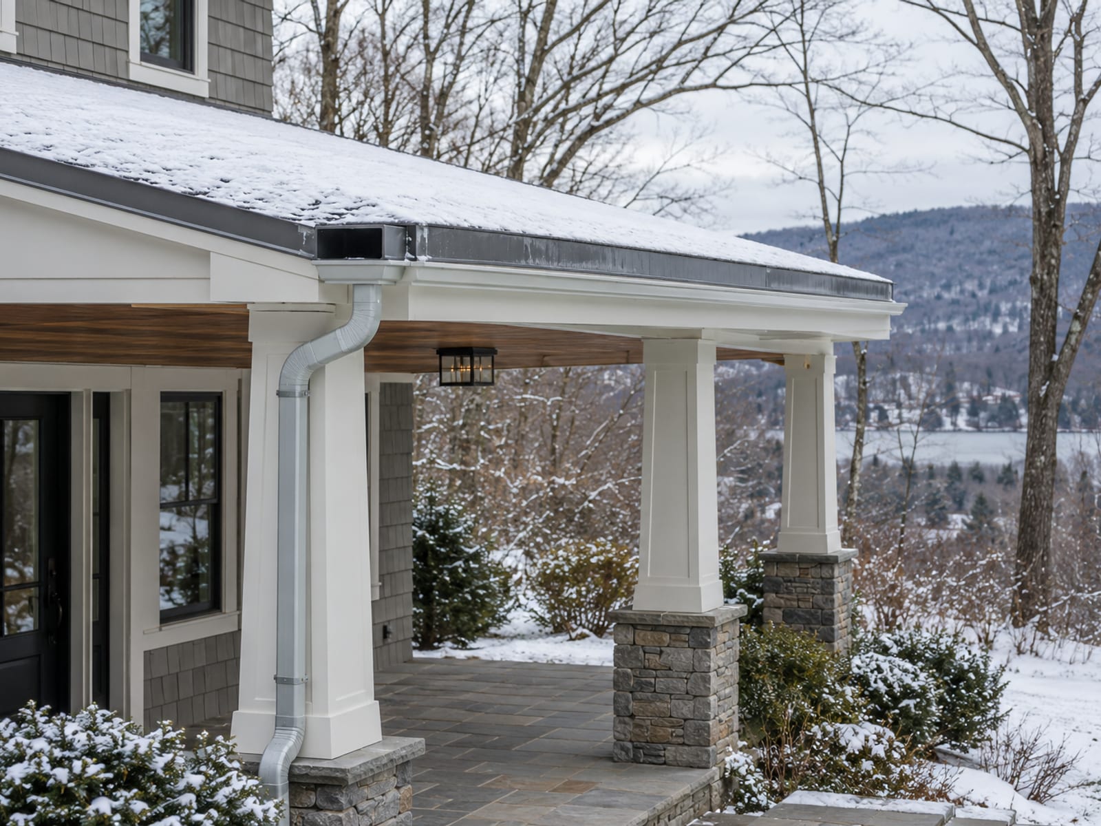Porch roof with a light dusting of snow, a clear metal scupper at the edge and a downspout, seen from the yard