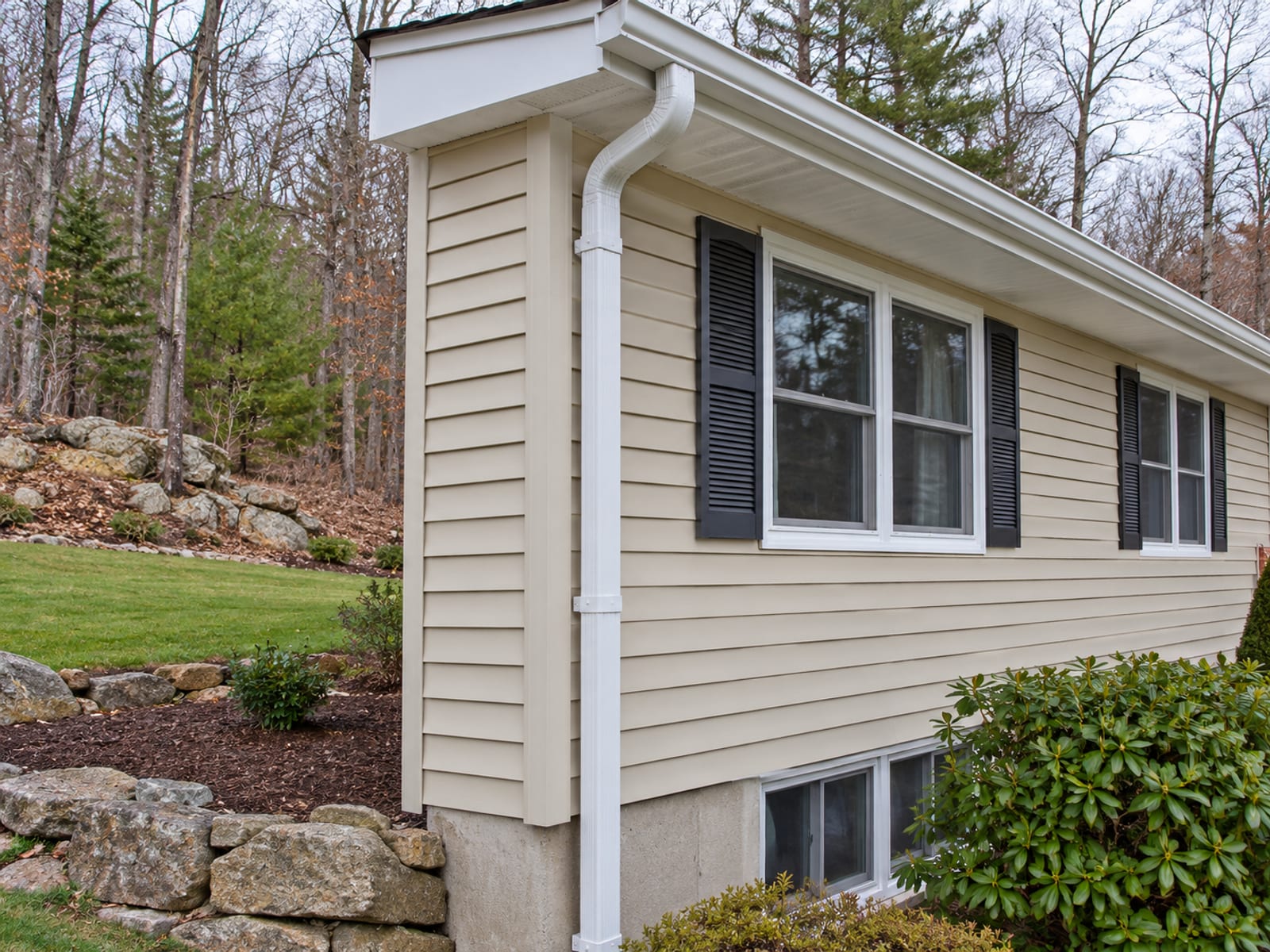 Finished white downspout fastened to beige vinyl siding with evenly spaced brackets, below a clean gutter outlet and straight fascia on a split-level house