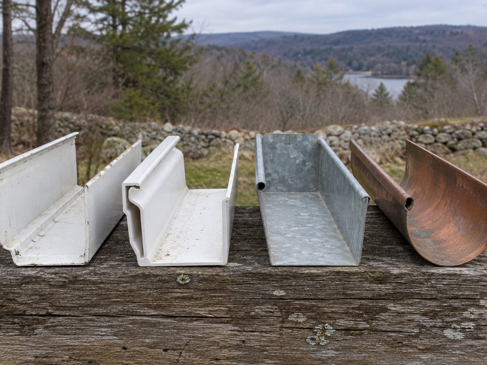 Four short gutter sections in white aluminum, white vinyl, gray galvanized steel and copper lined up on a weathered wooden plank