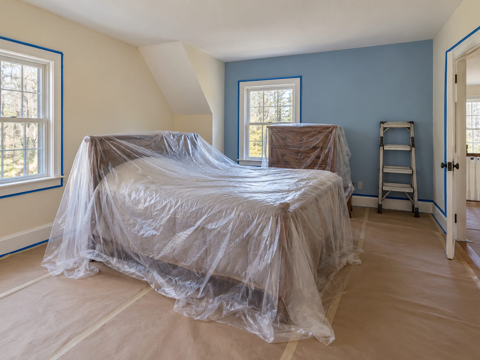 Bedroom with furniture gathered in the middle under plastic sheeting, floor covered in paper, one wall painted and the others still unpainted