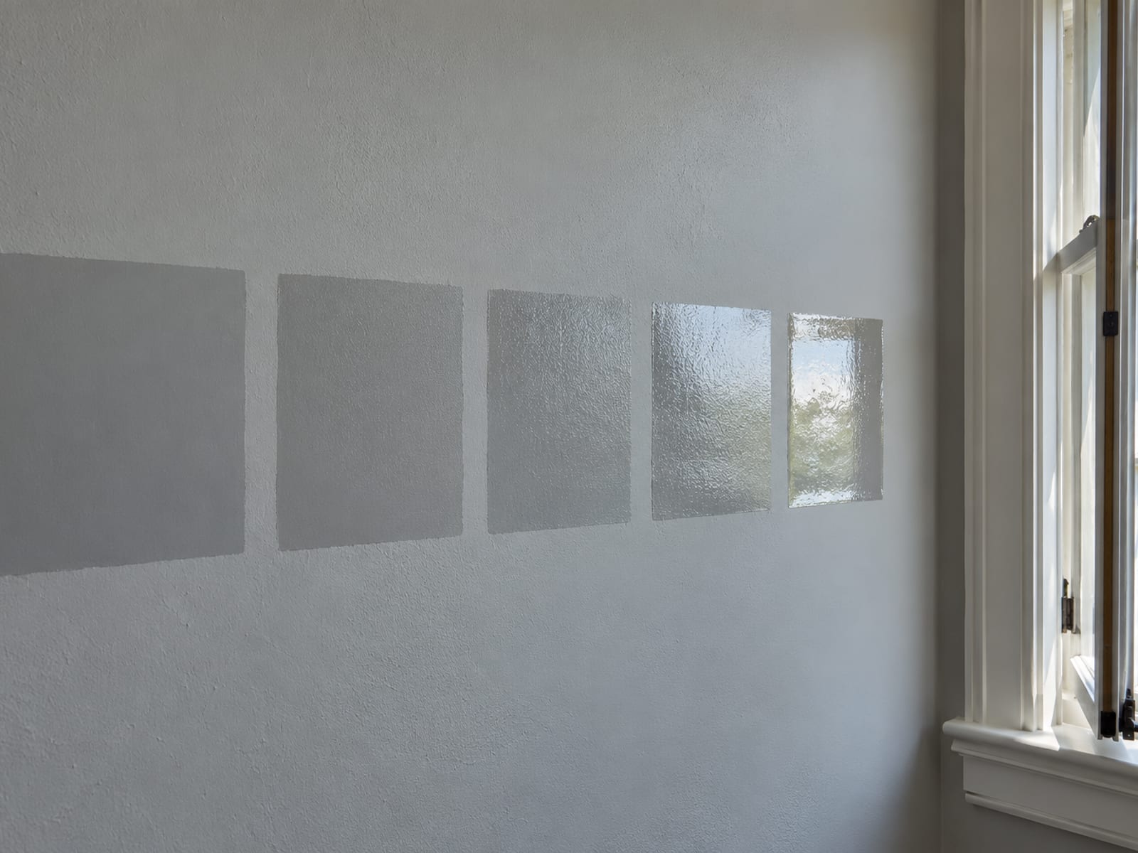 Painted wall with five square sample patches in sheens from flat to high gloss, catching window light differently beside a window