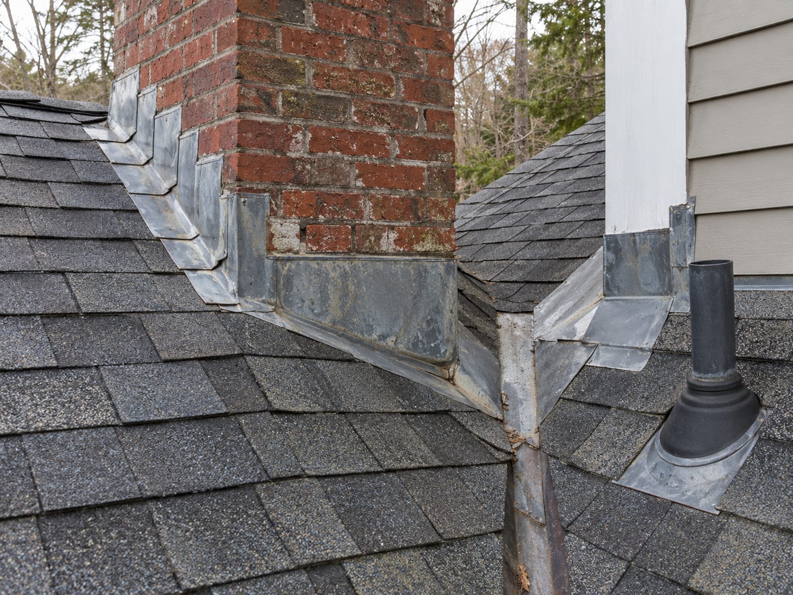 Roof corner where a brick chimney, a sidewall, a plumbing vent pipe and a valley meet, with visible metal flashing and asphalt shingles