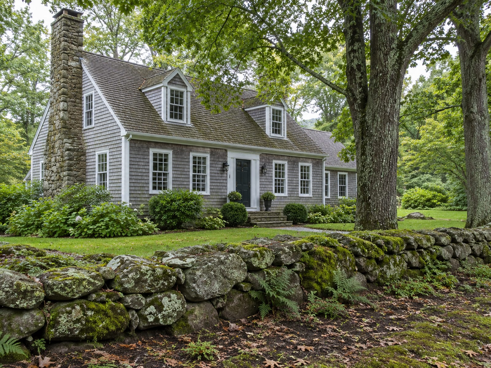 Weathered cedar shake Cape house behind a stone wall, shaded by tall oaks near a lake-country road