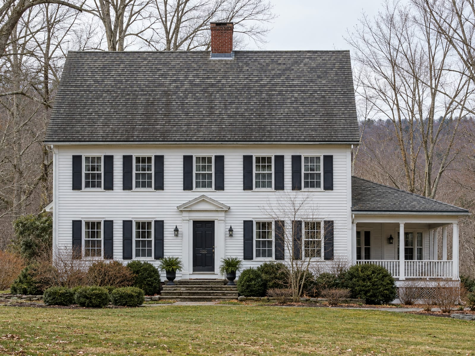Two-story colonial house with a steep gabled asphalt shingle roof, a brick chimney and a lower porch roof seen from across a winter street
