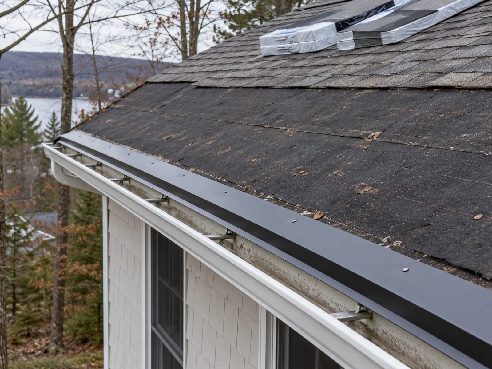 Roof edge with old shingles stripped back to underlayment, new drip edge along the eave and an aging gutter hanging in front of the fascia