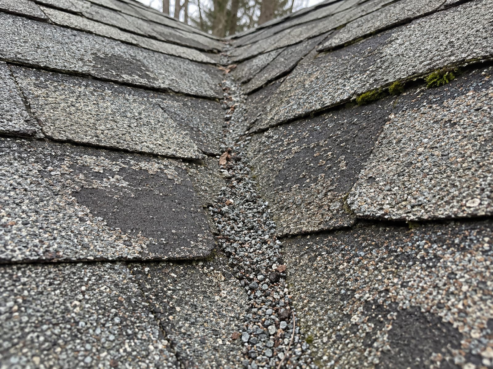 Close-up of old asphalt shingles with worn bare patches, collected loose granules and a moss edge near a roof valley