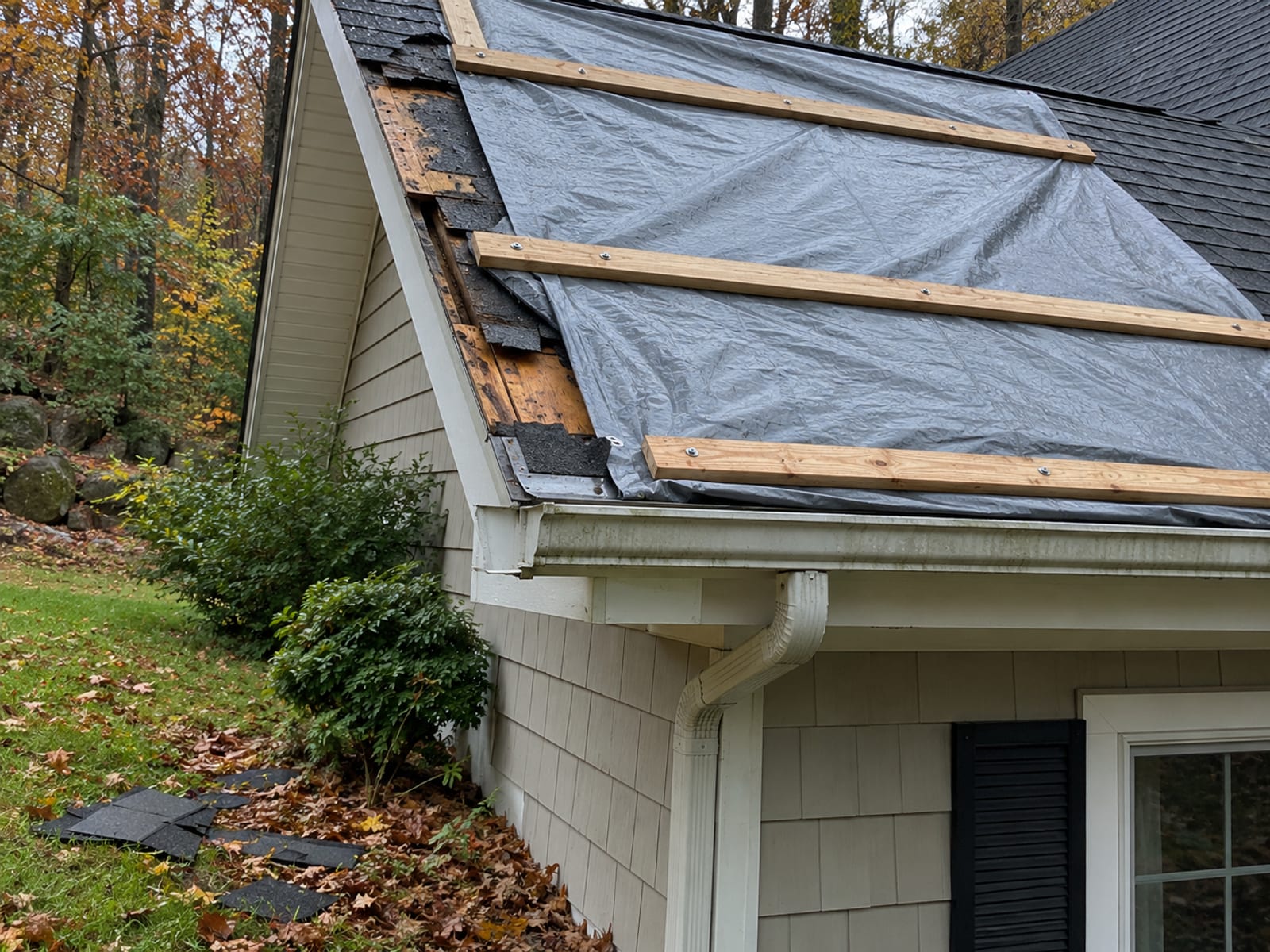 Gray heavy-duty tarp secured with wood battens over a damaged roof corner at the eave of a house, with a few fallen shingles on the lawn below