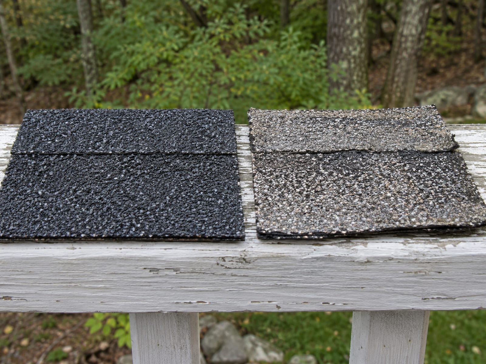 Close-up of a new charcoal shingle piece lying beside a faded weathered gray shingle piece on a wooden porch rail