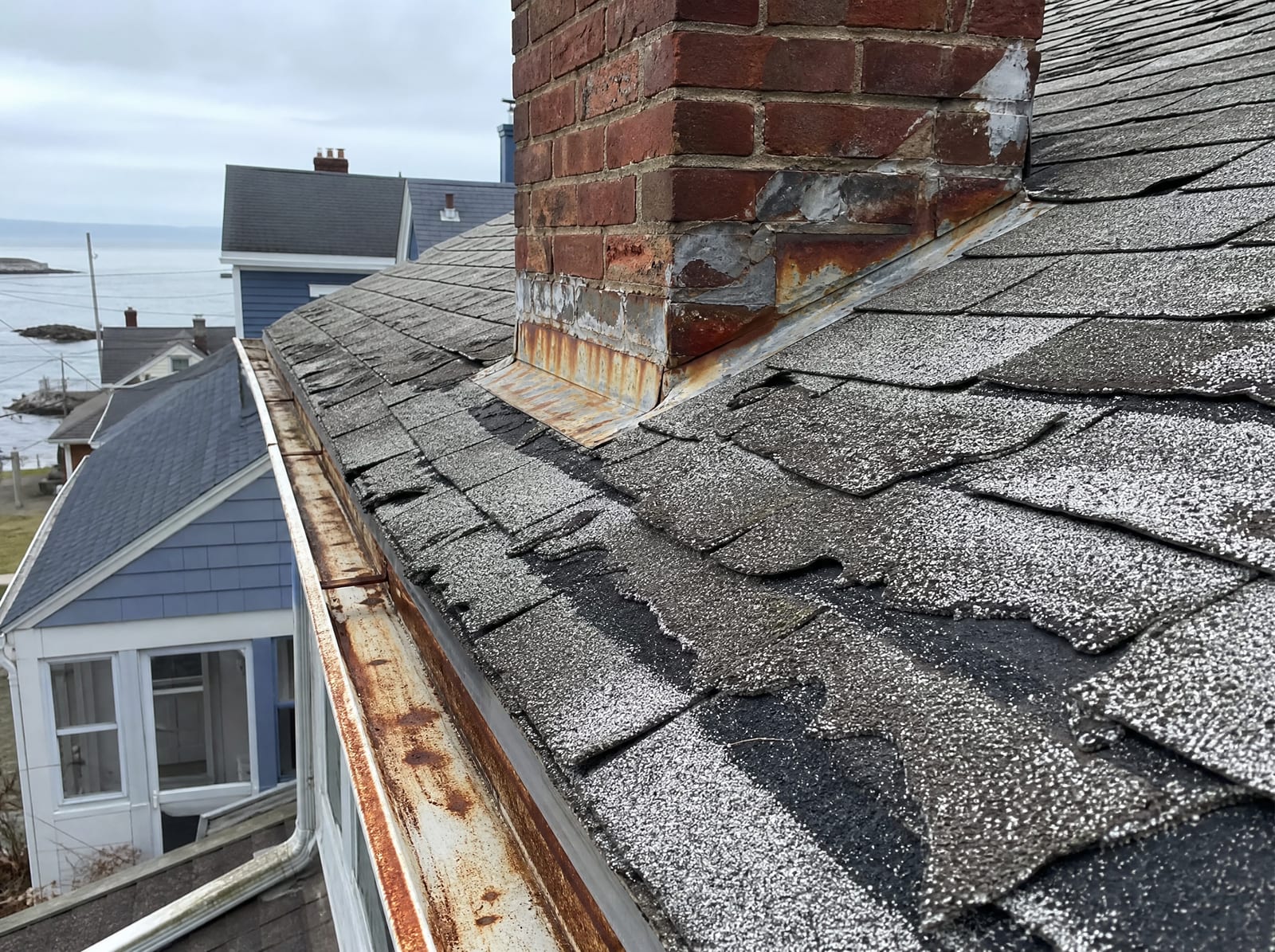 Aging asphalt shingle roof on an older coastal city home with granule loss, curling shingles, and salt-corroded rusted flashing