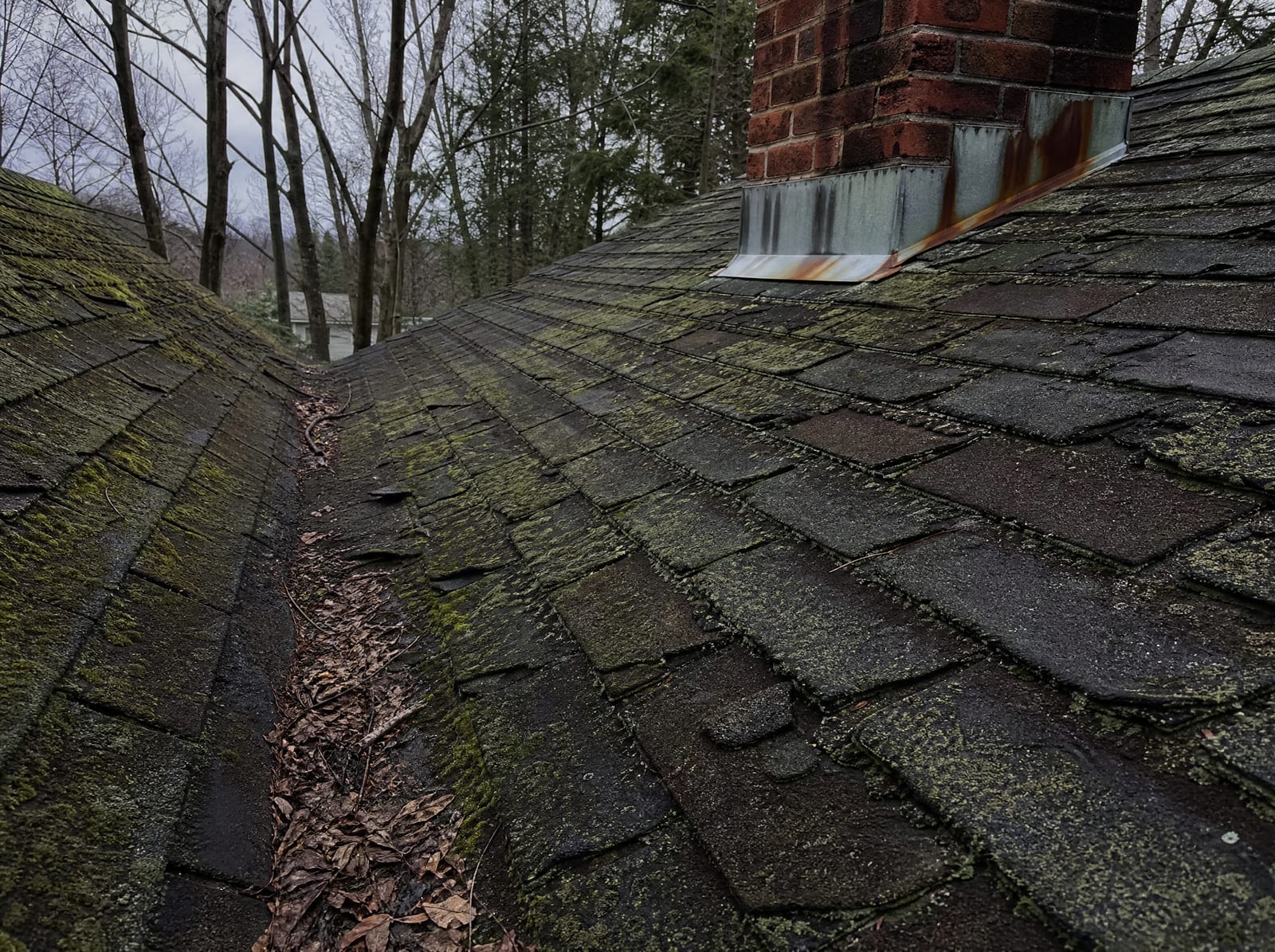 Aging asphalt shingle roof on an older Cheshire home with moss on a shaded slope, granule loss, curling shingles, and rust-streaked chimney flashing
