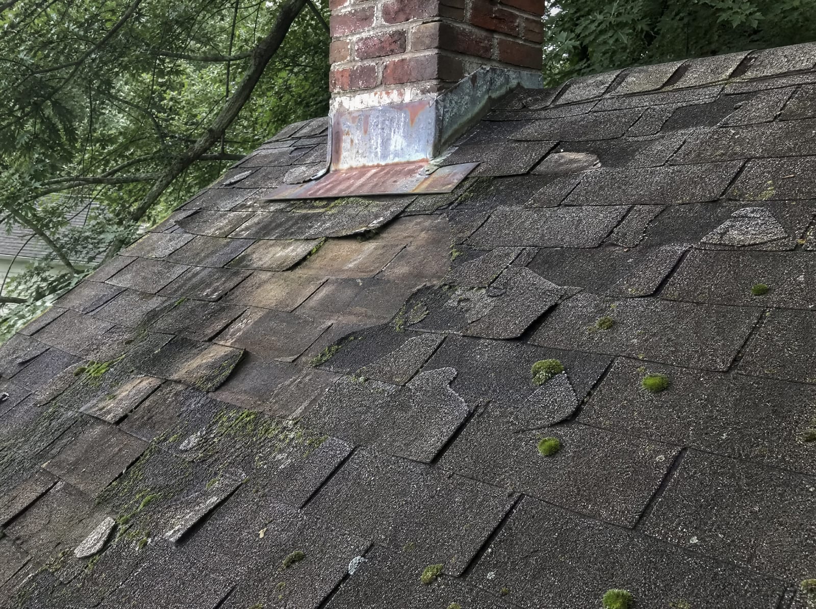 Aging asphalt shingle roof with widespread granule loss, curling and cracked shingles, green moss on a shaded slope, and worn chimney flashing