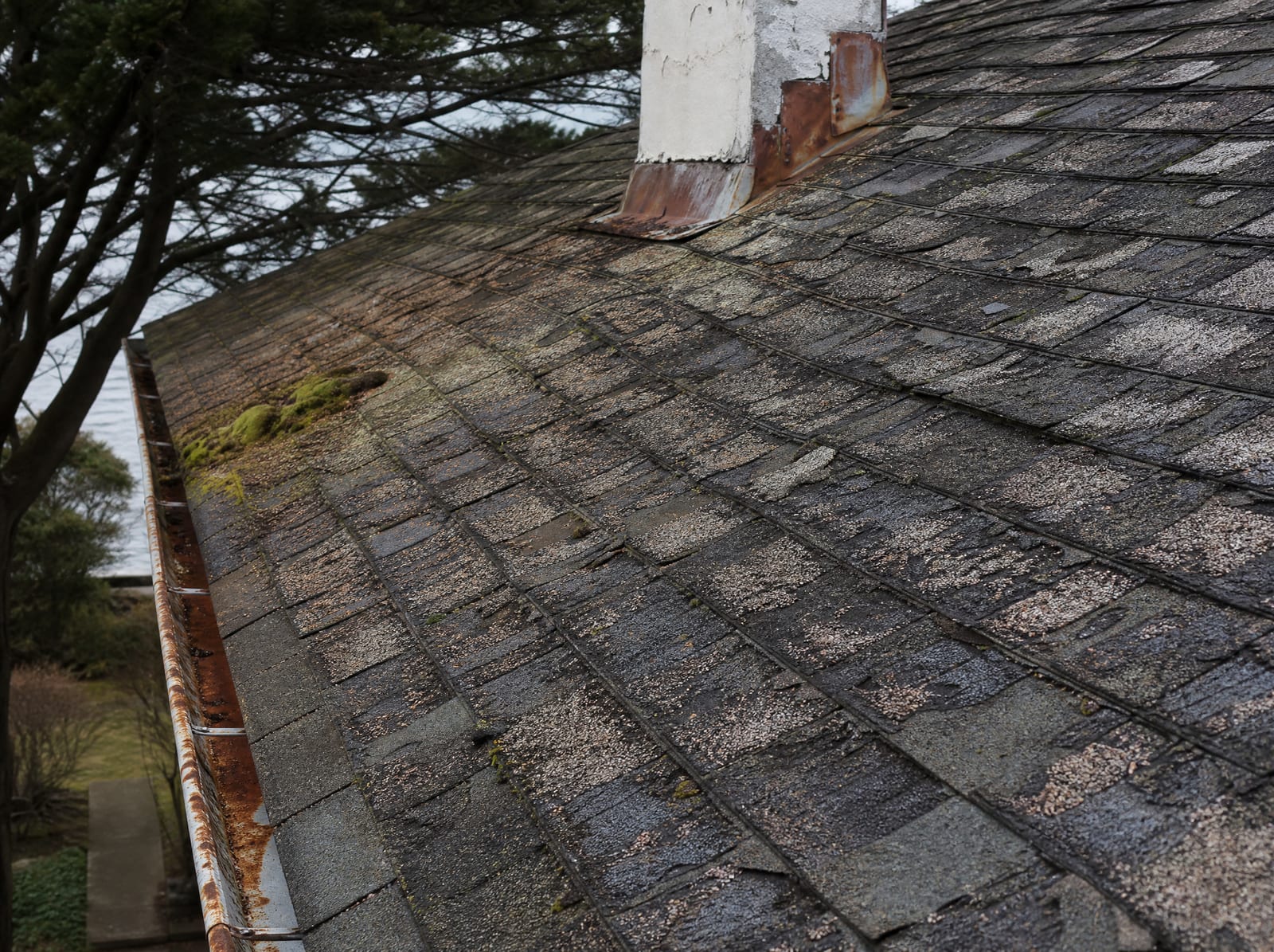 Aging asphalt shingle roof on an older coastal home with granule loss, curling shingles, moss, and salt-corroded flashing