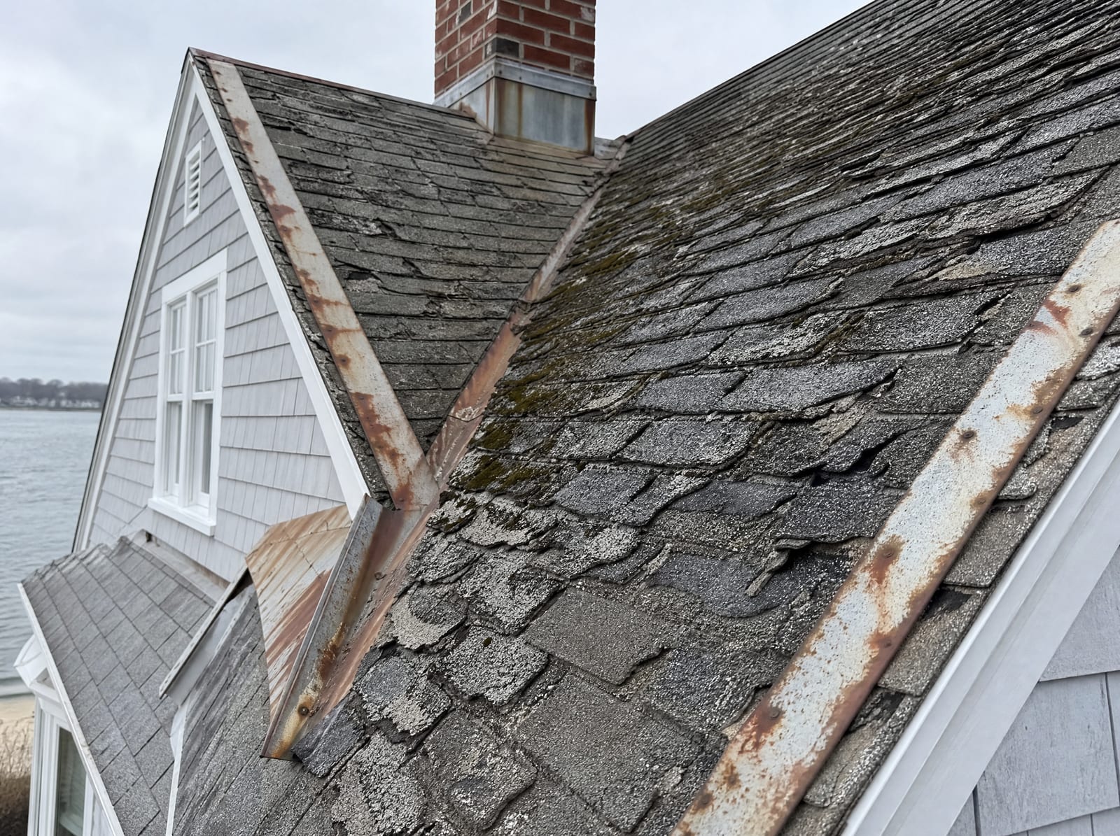 Aging asphalt shingle roof on an older coastal Milford Cape home near the shore with salt-corroded flashing, granule loss, and curling shingles