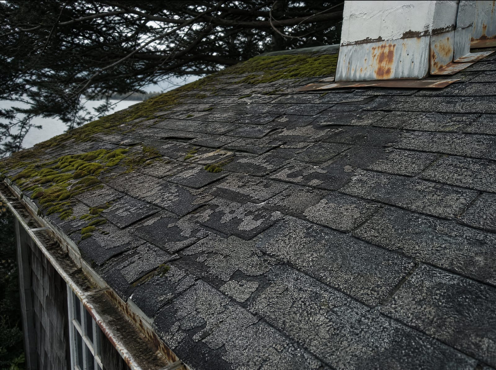 Aging asphalt shingle roof on an older coastal home with granule loss, curling shingles, moss, and salt-corroded flashing