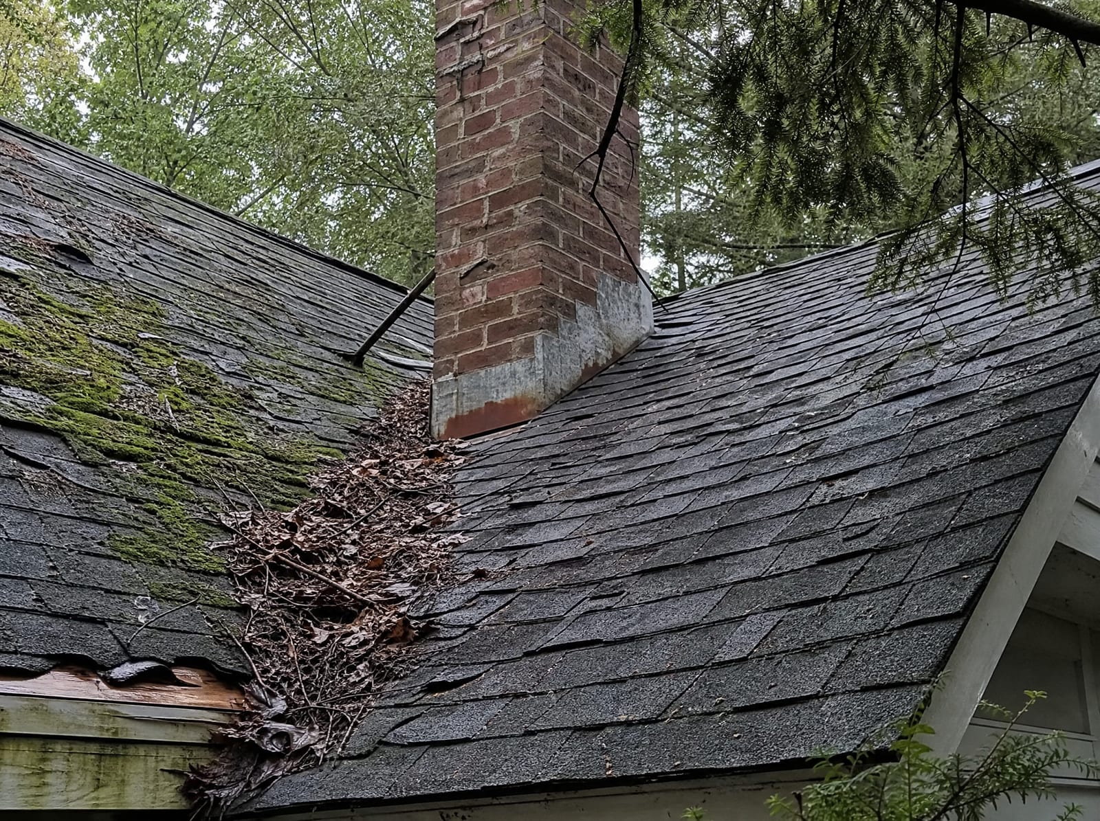 Aging asphalt shingle roof on an older Oxford home with moss on a shaded slope, granule loss, curling shingles, and rust-streaked chimney flashing