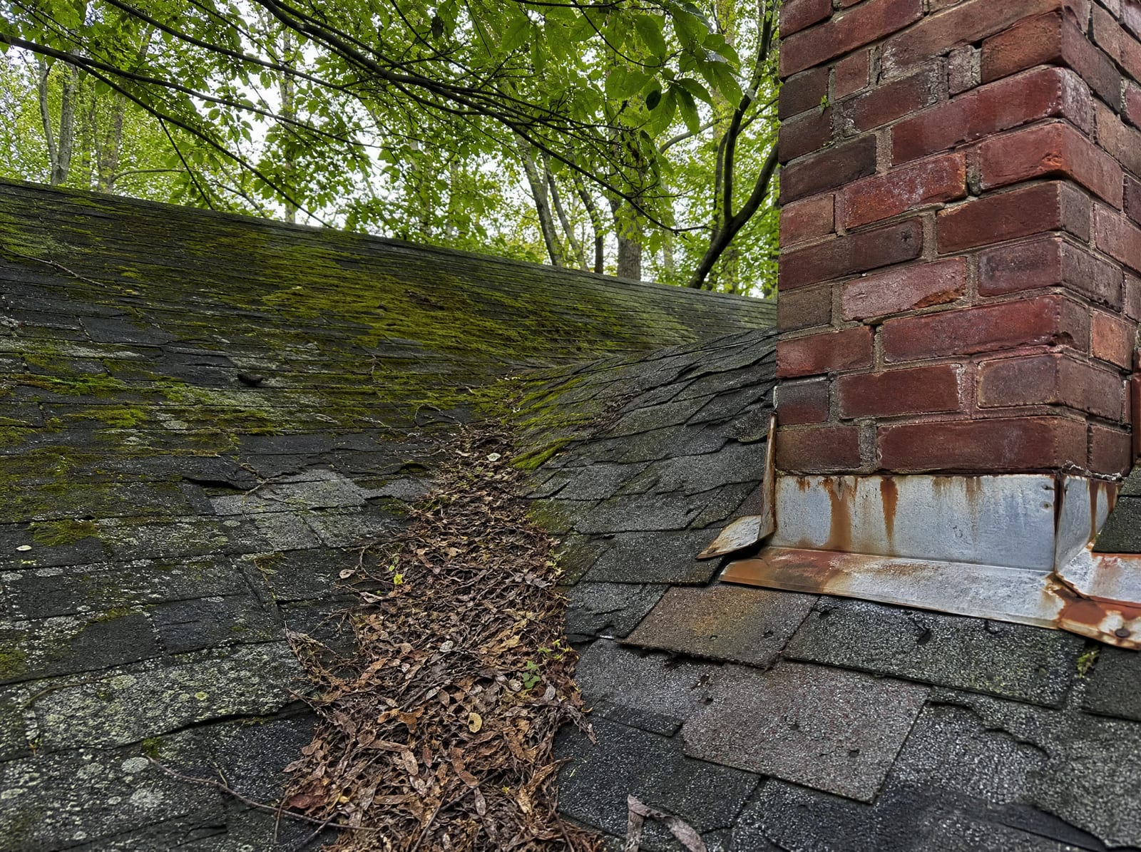 Aging asphalt shingle roof on an older Southbury home with moss on a shaded slope, granule loss, curling shingles, and rust-streaked chimney flashing