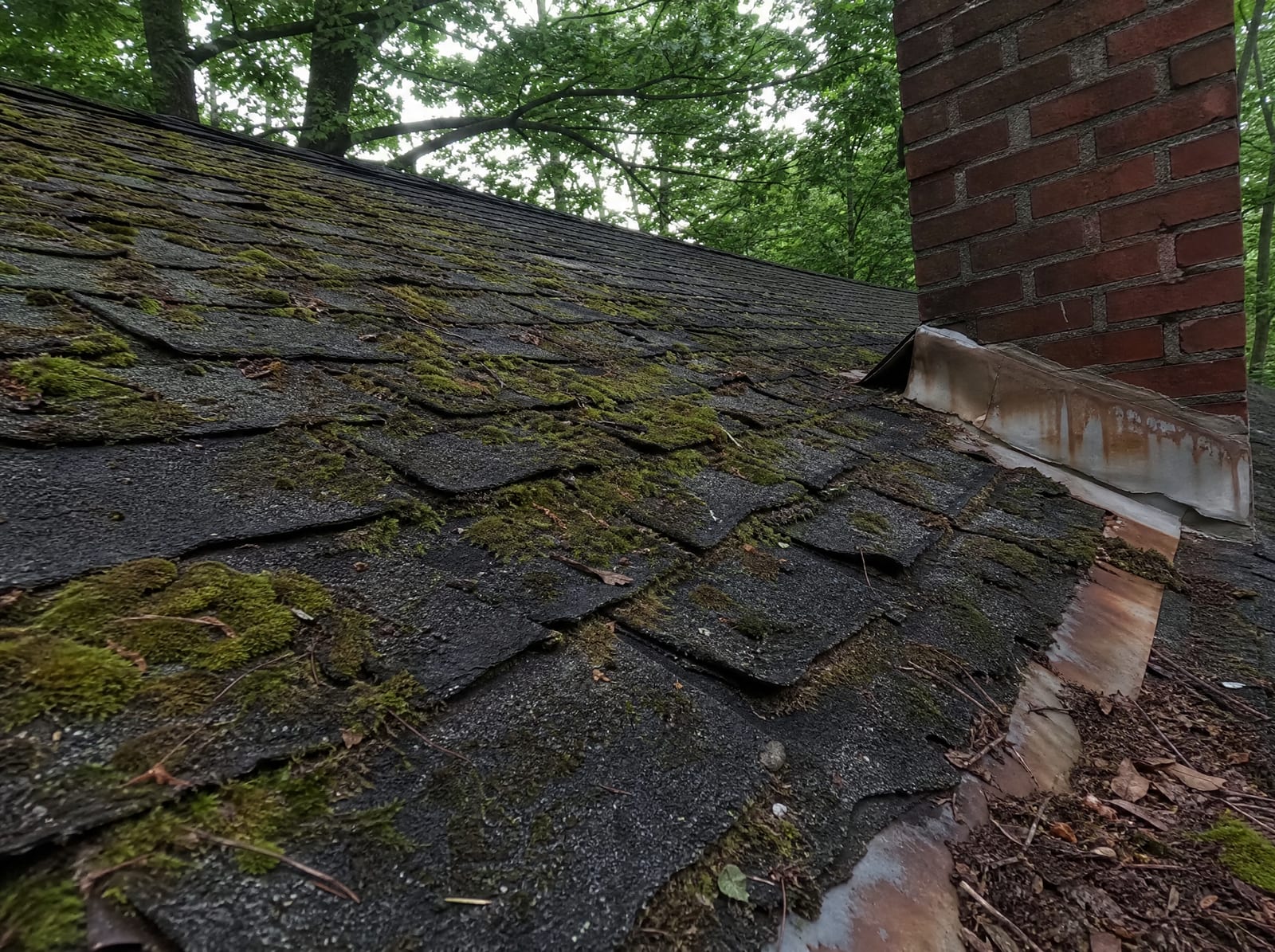 Aging asphalt shingle roof on an older Watertown home with moss on a shaded north slope, granule loss, curling shingles, and rust-streaked chimney flashing