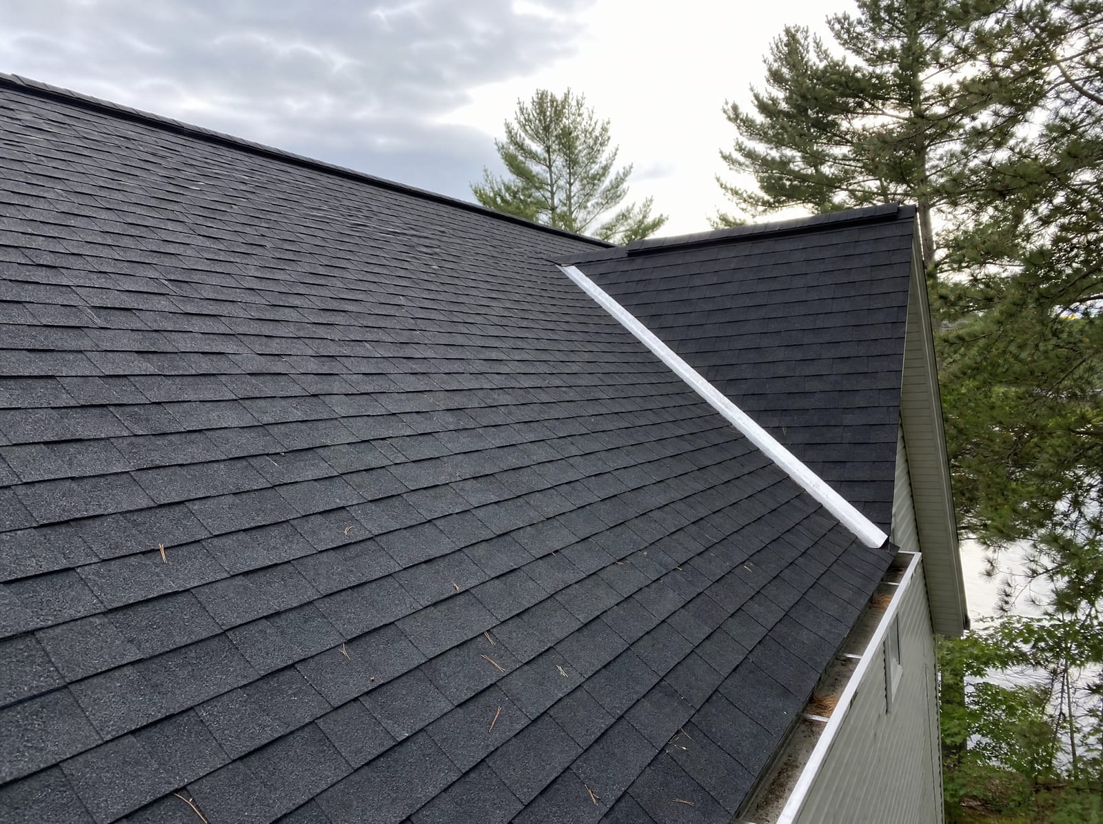 Freshly completed dark architectural asphalt shingle roof on a wooded lake-area home with new metal valley flashing and a continuous ridge vent