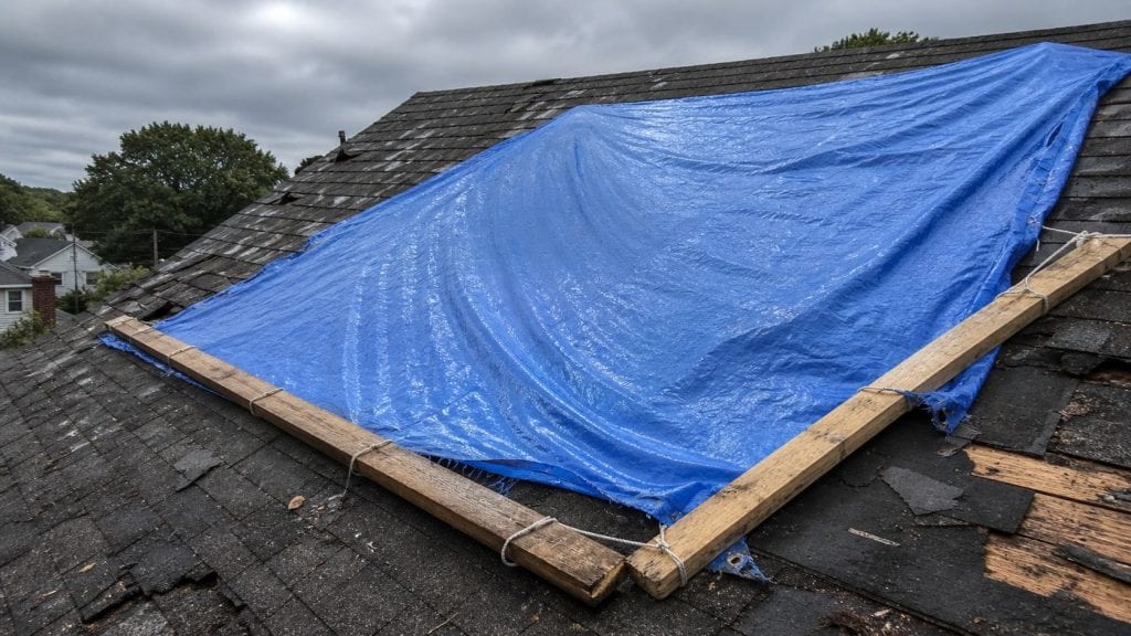 A bright blue polyethylene tarp stretched tightly across an exposed section of a residential roof, secured with weighted boards along its edges, covering missing shingles and exposed underlayment