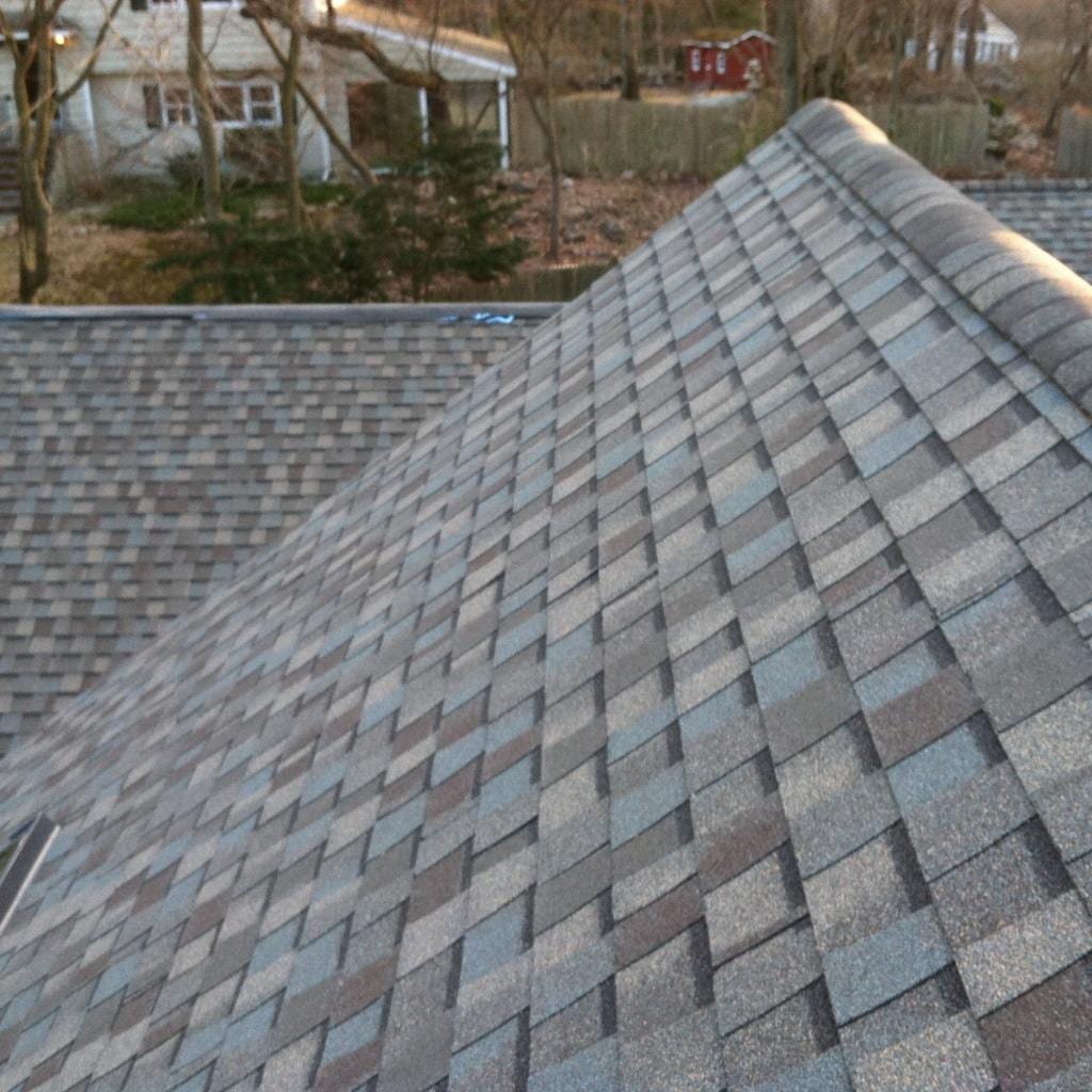 Close-up view of a newly installed multi-tonal gray and brown architectural asphalt shingle roof with a hip ridge line, overlooking a suburban Connecticut backyard with bare trees, a white house, and a red outbuilding in the background