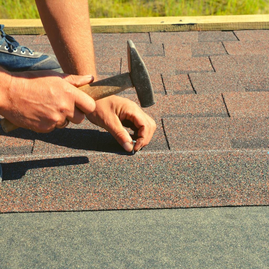 A roofer kneeling on a roof hammering a nail into red and gray asphalt shingles along the roof edge in daylight