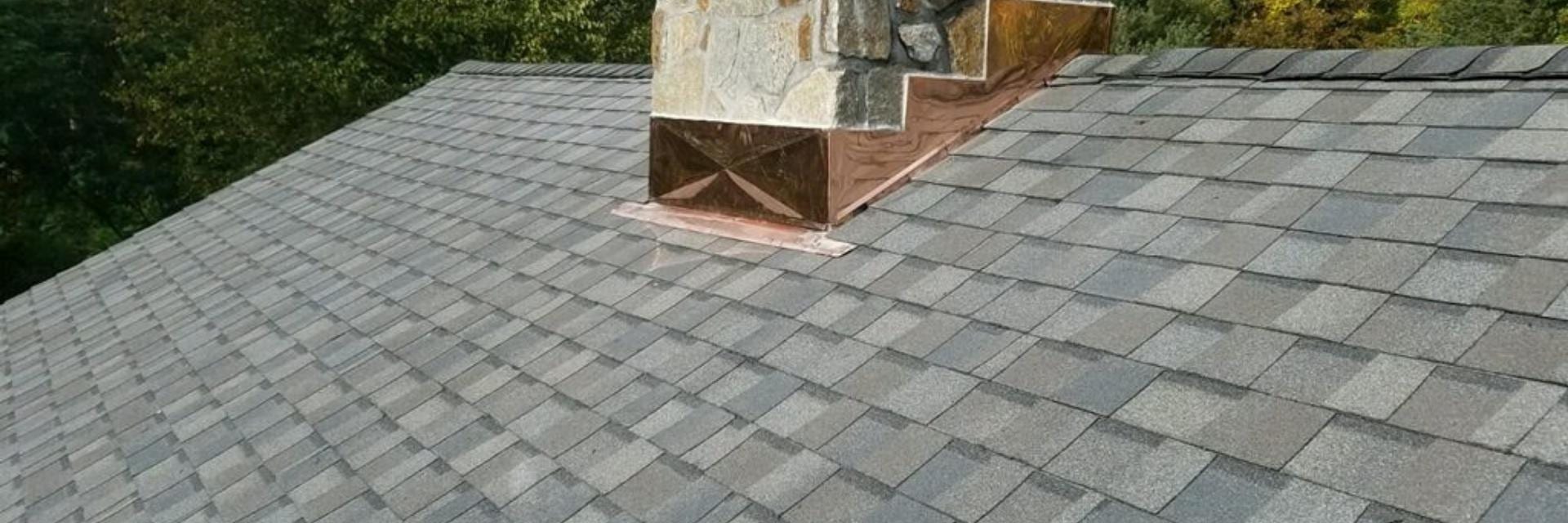 Gray asphalt shingle roof with a stone chimney wrapped in new copper flashing, surrounded by green trees in the background