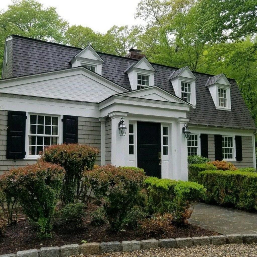 Gray shingled Cape Cod style home with white trim black shutters dormers and black front door surrounded by green shrubs and trees