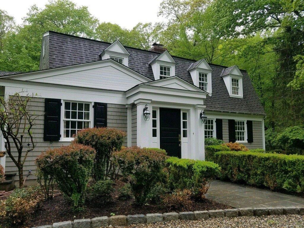 Cape Cod style home with gray cedar shake siding, dark asphalt shingle roof with three dormers, black shutters and front door, white trim portico entry, surrounded by mature trees and manicured shrubs