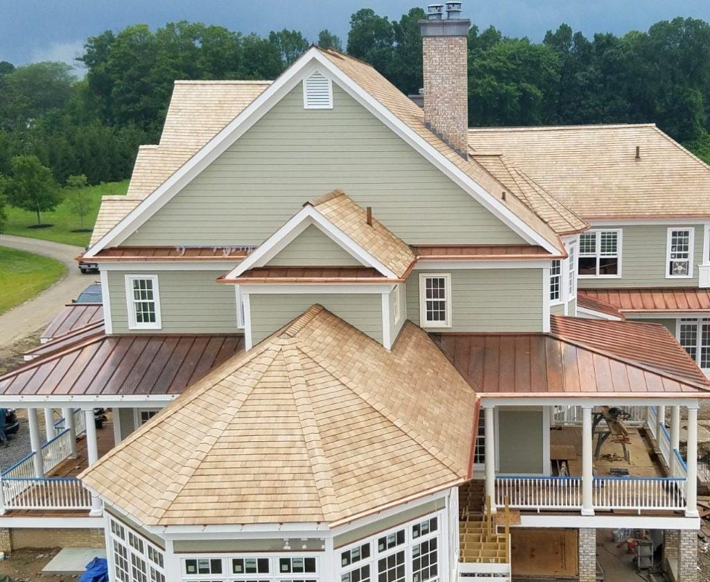 Aerial view of a large multi-gabled home under construction featuring fresh cedar shake shingles on pitched roofs and standing seam copper metal roofing on porch sections with white trim and sage green siding