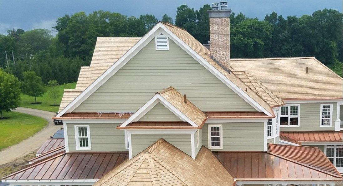Aerial view of a large multi-gabled home with newly installed cedar shake shingles and copper standing seam metal roof sections, sage green horizontal siding, white trim, brick chimney, and multiple dormers surrounded by dense green trees in Connecticut