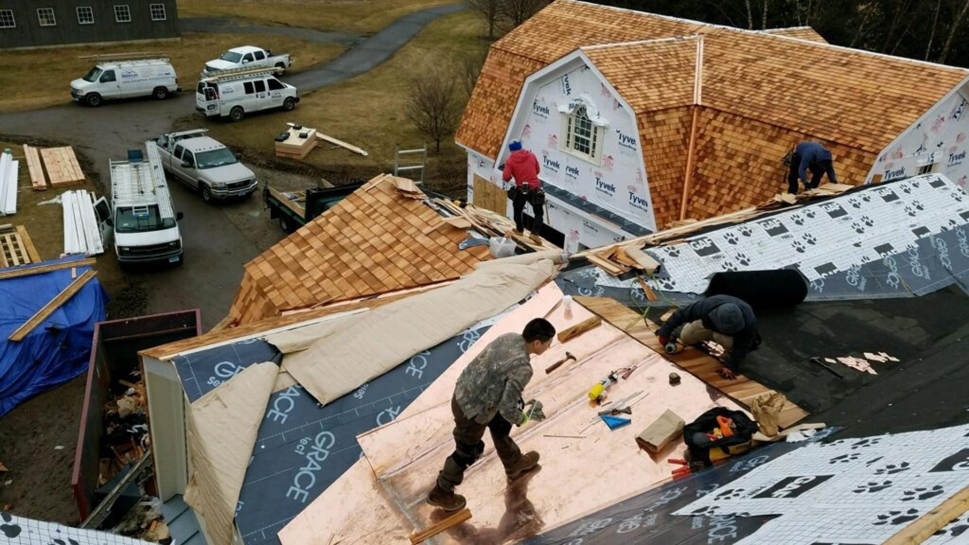 Aerial view of multiple roofers installing cedar shake shingles on a large residential roof with Grace ice and water shield underlayment visible, Tyvek house wrap on adjacent gable, and contractor work trucks parked on gravel driveway below