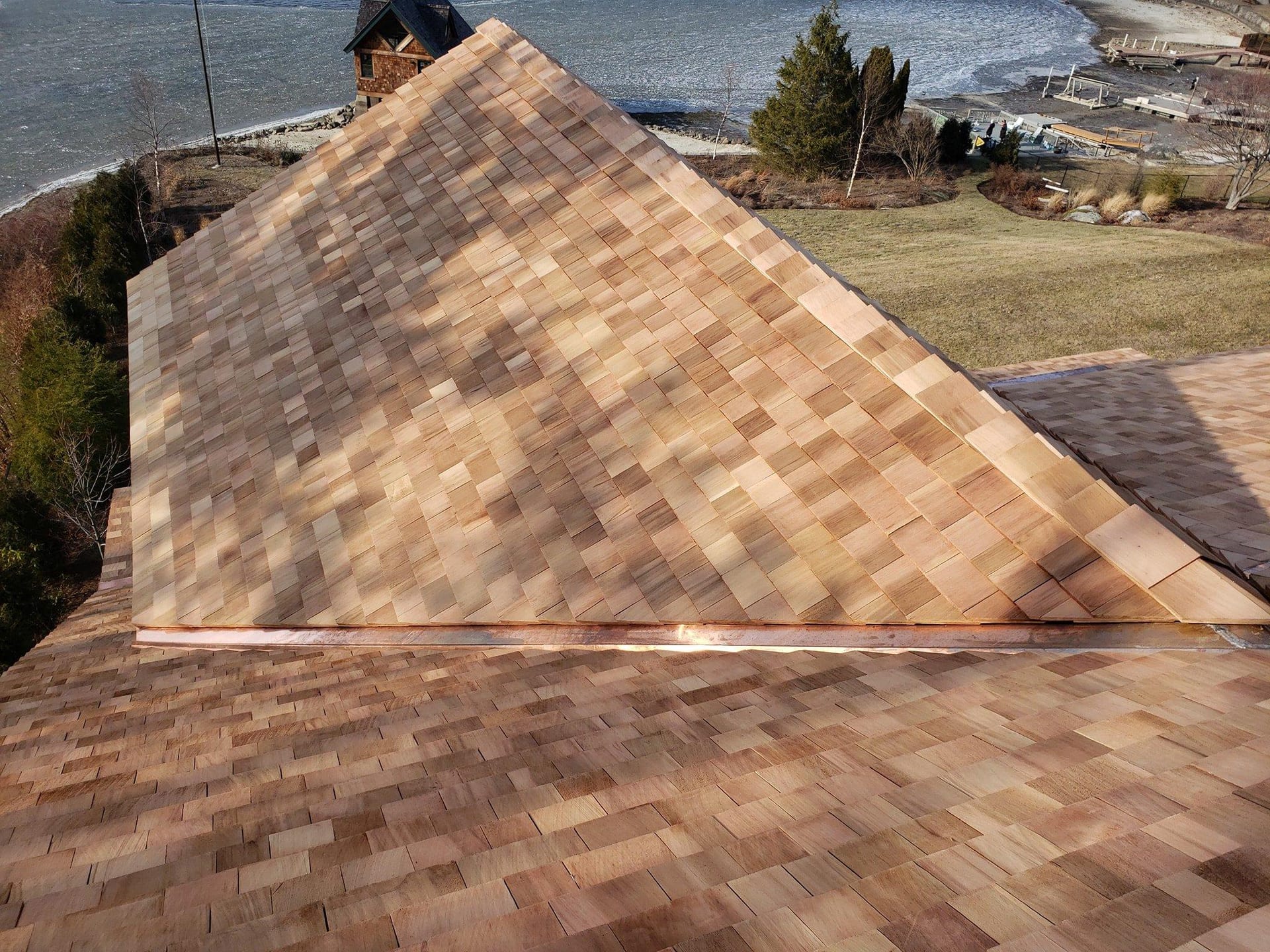 Freshly installed cedar wood shake shingles covering a steep-pitch hip roof with a copper valley flashing strip, overlooking a coastal lawn and shoreline with a neighboring cottage and dock in the background