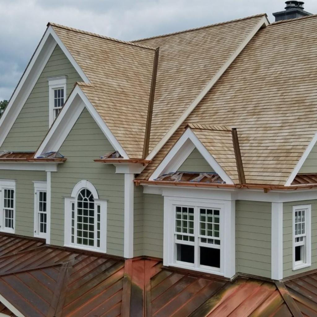 Aerial view of a sage green colonial house with new cedar shake shingles, copper flashing at valleys and dormers, multiple gabled dormers with white trim, arched and grid windows, and a copper standing seam lower roof section under overcast skies