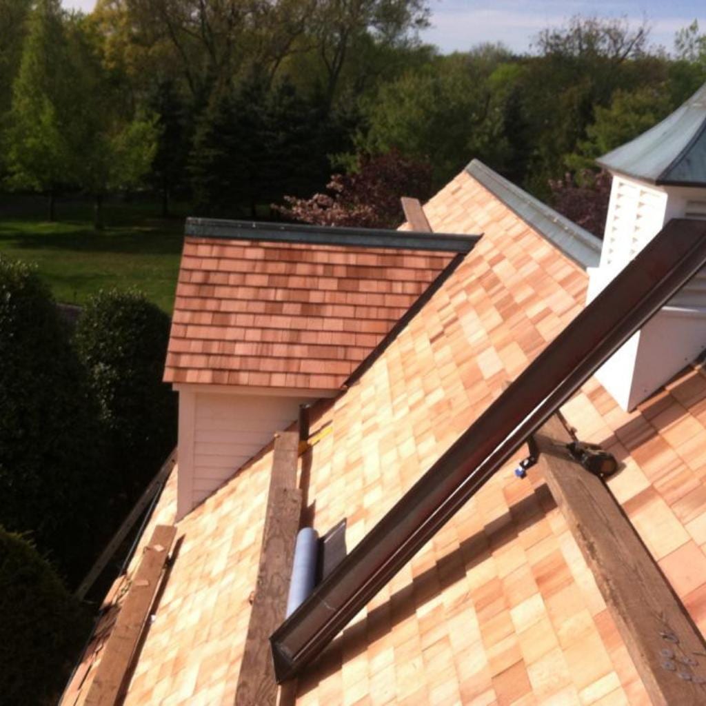 Aerial view of freshly installed cedar shake shingles covering multiple roof planes with dormers, a white cupola with copper cap, rain gutters, and surrounding green trees and lawn