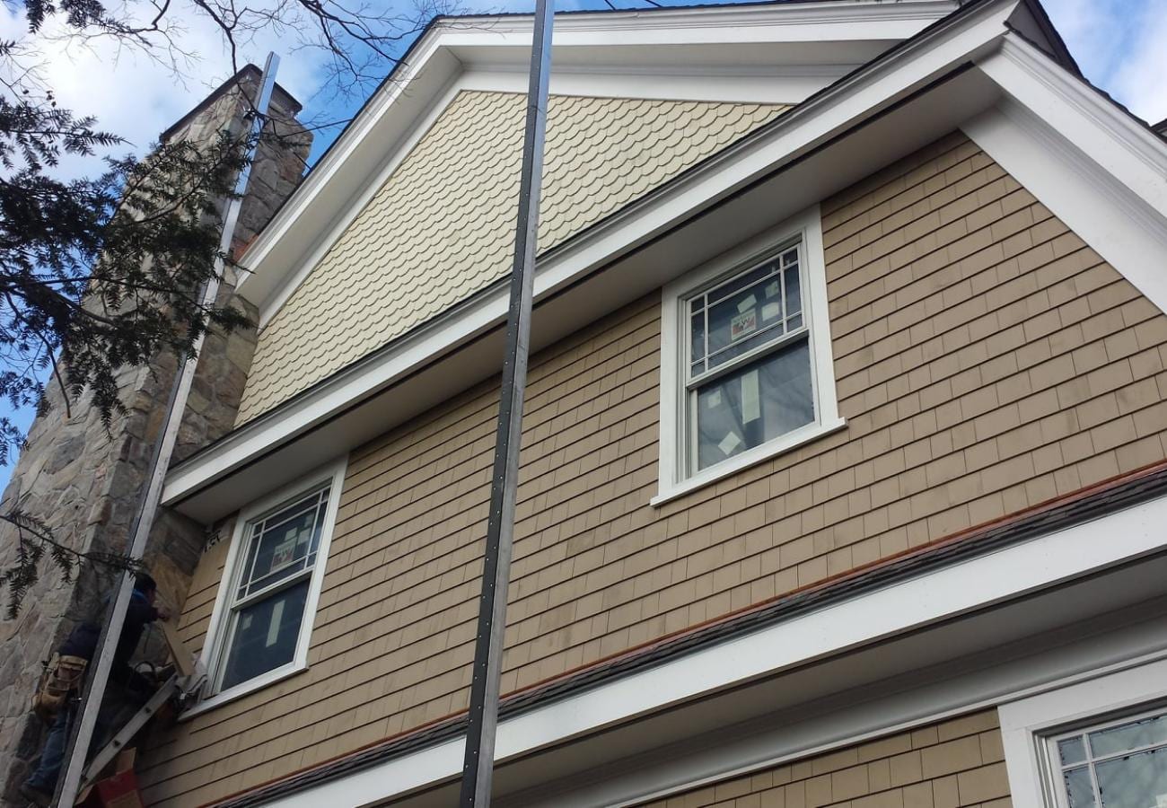 Upward angle view of a two-story home exterior featuring newly installed tan cedar shake lap siding, scalloped shingle accents on the upper gable, white trim around double-hung windows with grilles, a stone chimney on the left, and a metal scaffold pole in the foreground