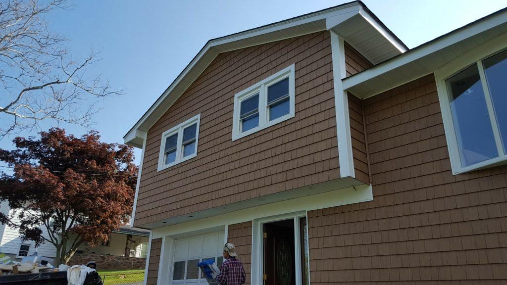 Worker in plaid shirt and cap installing cedar shake vinyl siding on a two-story home with white trim windows and an attached garage under a clear blue sky