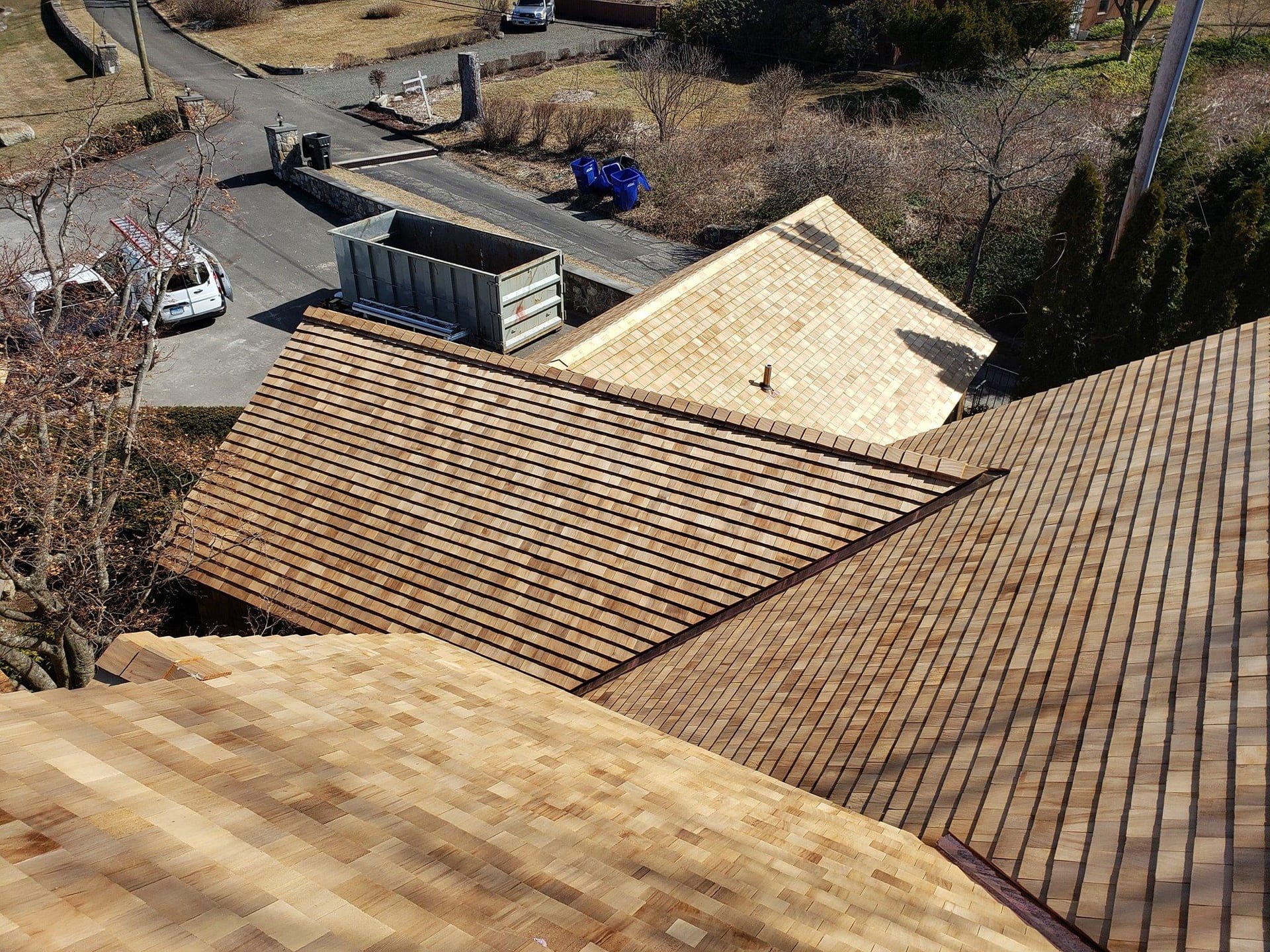 Aerial view of a multi-section cedar wood shake roof installation on a residential home with a dumpster in the driveway below, surrounded by bare winter trees and a neighborhood street