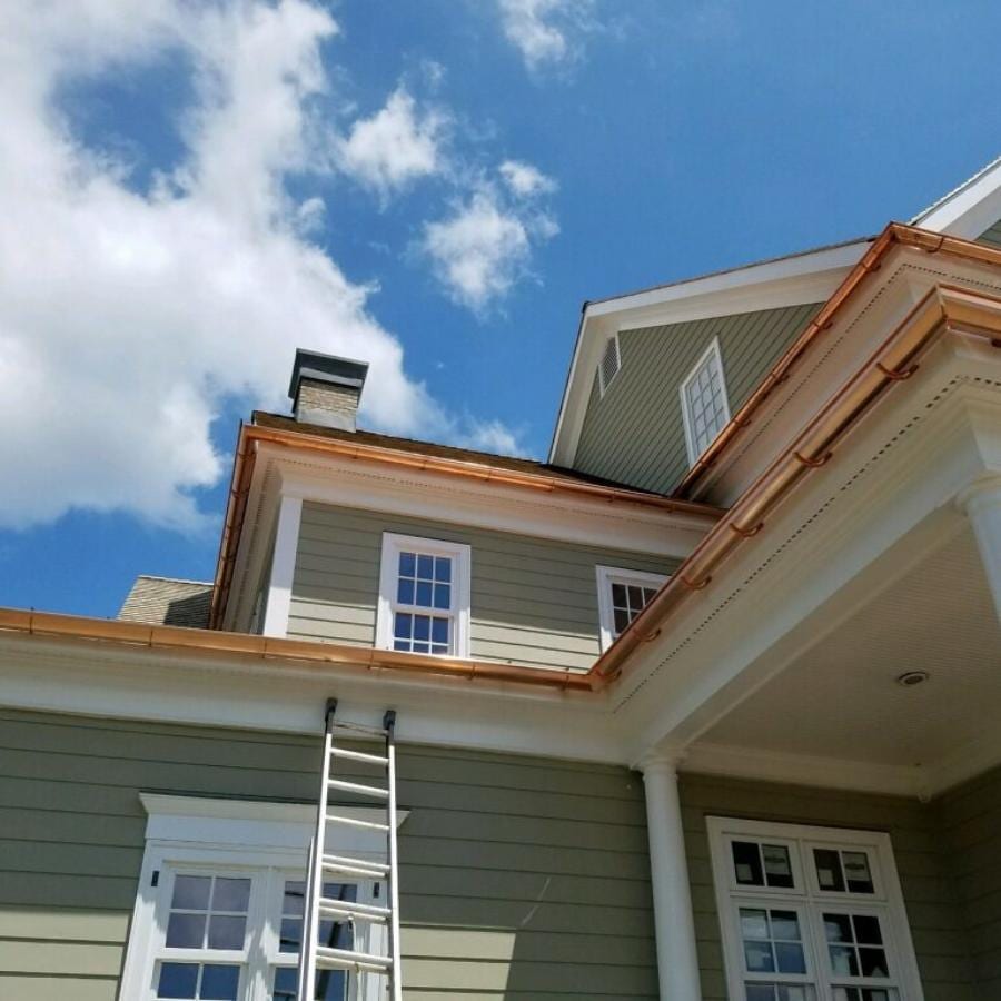 Newly installed copper gutters and downspouts along the roofline of a two-story sage green colonial home with white trim, a ladder leaning against the first-floor eave, and a blue sky with scattered clouds above