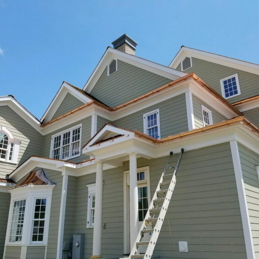 Newly installed copper gutters and downspouts on a large two-story sage green lap siding home with white trim, dormers, and a chimney, with an aluminum extension ladder leaning against the front facade
