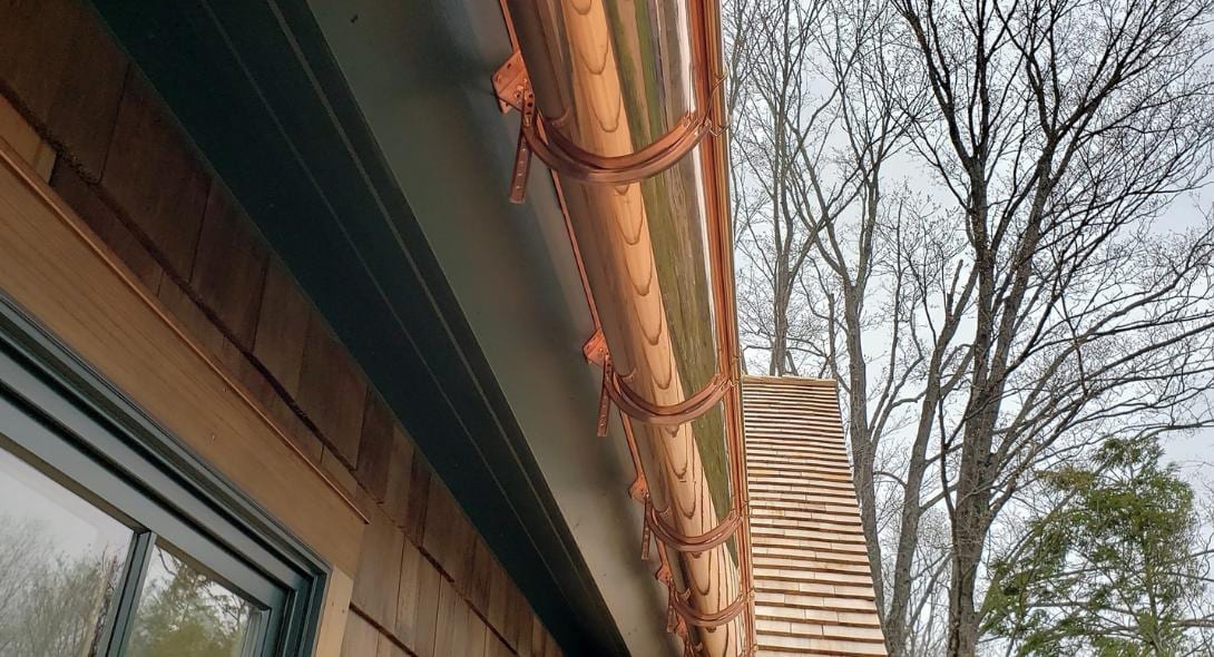 Newly installed shiny copper half-round gutters with copper brackets mounted along a dark soffit above cedar shake siding on a Connecticut home, with bare deciduous trees visible in the background