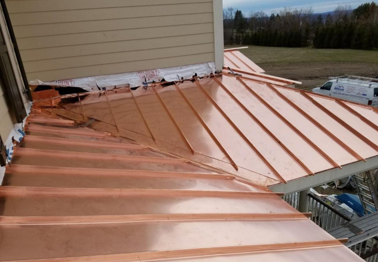 Newly installed copper standing seam metal roof panels fanning outward from a hip corner on a residential home, with DuPont underlayment visible along the wall flashing line and a contractor van parked in the background