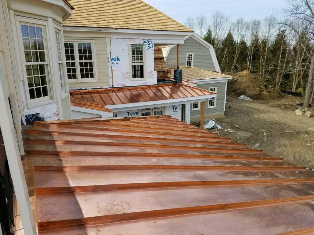 Freshly installed copper standing seam roof panels on a residential addition under construction, with Tyvek house wrap visible on adjacent walls and cedar shake roofing on the main structure in the background