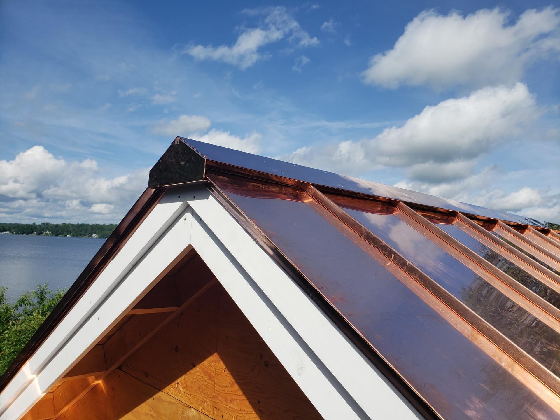 Newly installed copper standing seam roof panels on a gabled structure with white trim fascia boards, overlooking a lake and tree-lined shoreline under a partly cloudy blue sky