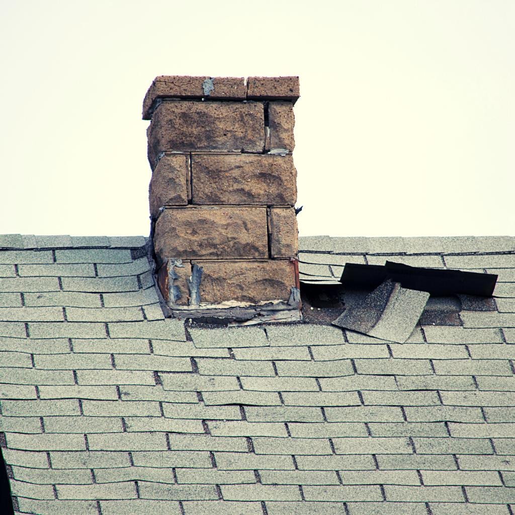 Weathered brick chimney with crumbling mortar and missing flashing sitting on a gray asphalt shingle roof with displaced shingles nearby