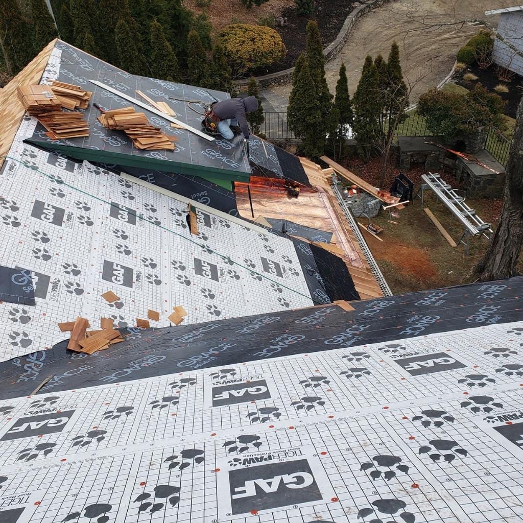 Roofer working on steep pitched roof covered with GAF Tiger Paw underlayment and Grace Ice and Water Shield, with cedar shake shingles stacked nearby and a residential yard visible below