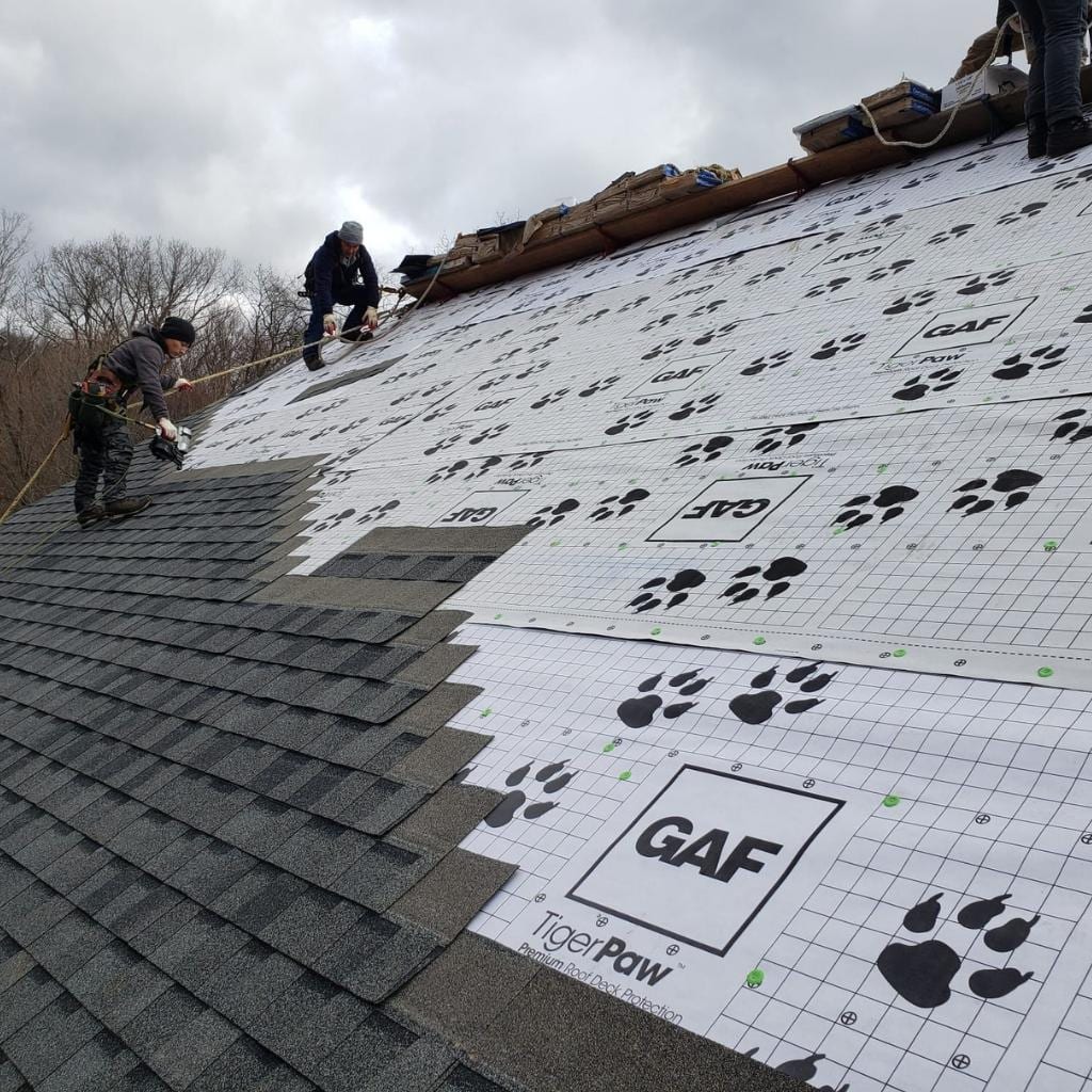 Two roofers wearing safety harnesses install GAF TigerPaw synthetic underlayment on a steep residential roof slope while gray architectural shingles are partially laid on the lower section