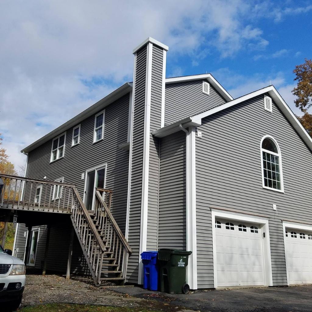 Two-story house with gray horizontal vinyl siding, white trim, arched window, exterior wood staircase leading to upper deck, two white garage doors, blue and green trash bins, and a partial view of a white SUV on a paved driveway during autumn