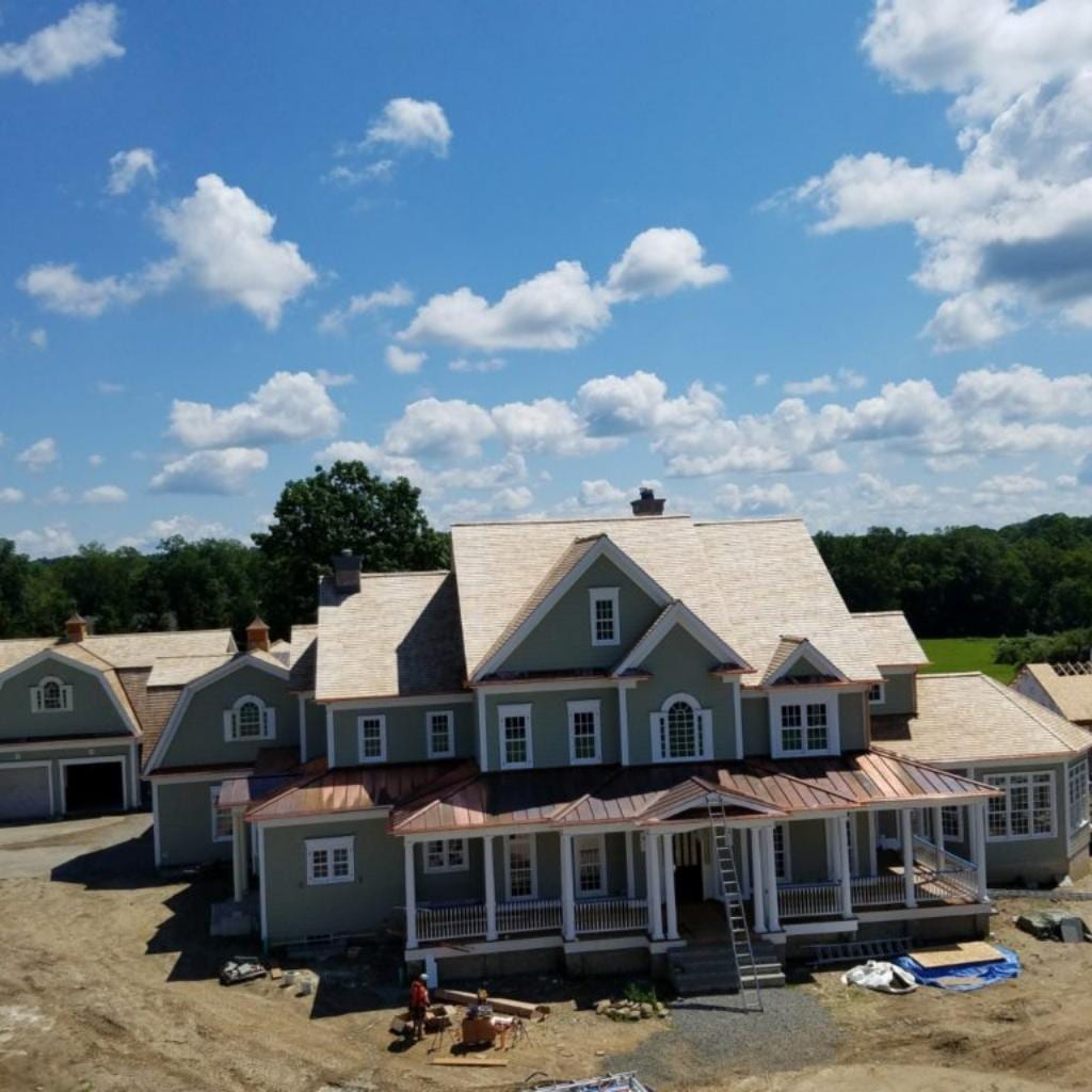 Aerial view of a large multi-wing estate home under construction in Connecticut featuring light cedar shake shingles, copper standing seam accent roofing on porches and dormers, gray siding with white trim, a wraparound front porch with columns, a detached garage with gambrel roof, a ladder leaning against the front facade, construction workers and materials on bare dirt grounds, surrounded by green trees under a partly cloudy blue sky