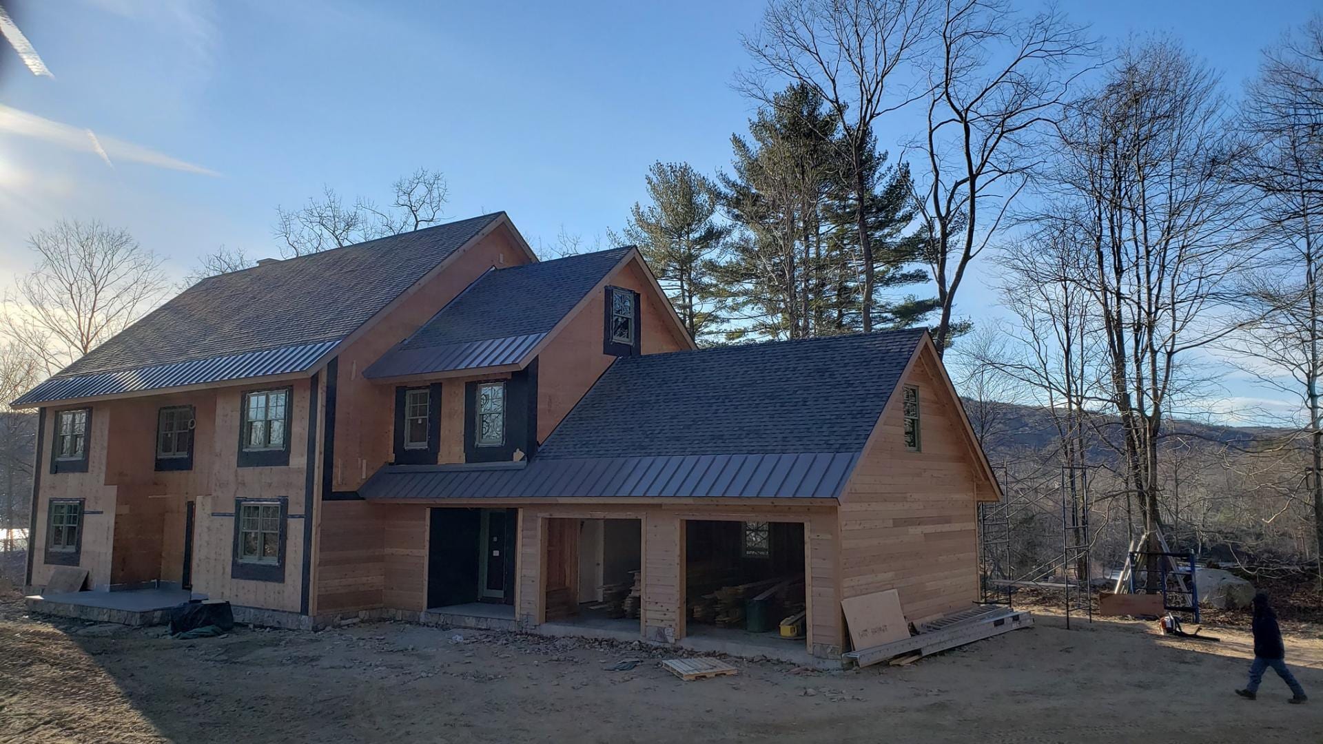 Two-story new construction home with mixed dark asphalt shingle and standing seam metal roofing, natural cedar wood siding partially installed, open three-car garage bays, scaffolding on right side, building materials on ground, surrounded by bare deciduous trees and evergreens with hills in background