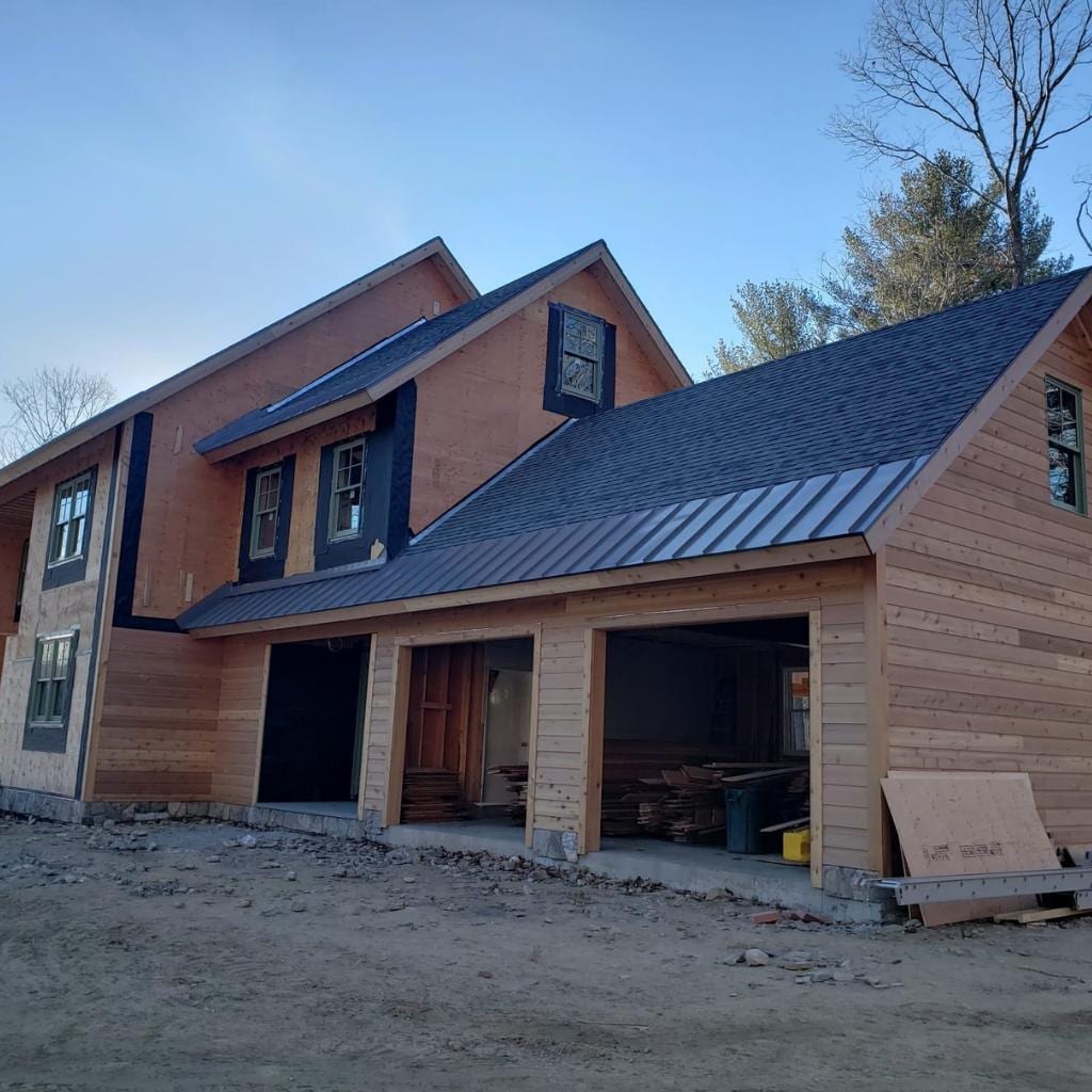 Two-story home under construction with natural cedar wood siding partially installed, dark asphalt shingles on the main roof, standing seam metal roof on the garage addition, and open garage bays storing lumber and building materials on a dirt lot
